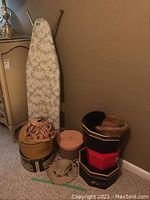 Photo showing the four hats stacked inside and beside six different collectible hat boxes, along with a floral patterned ironing board standing against the wall.