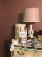 Wide view showing lamp with floral designs, box, night light, heart-shaped item, sunglasses, and pedestal container on a green table