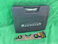Closed black plastic case with Mastercraft logo, Stanley Fatmax level placed in front on green fabric.
