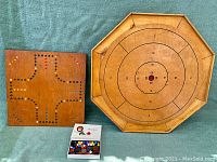 Wooden crokinole board is octagonal with markings for scoring and concentric score zones from 5 to 20 points; wooden cribbage board with colored peg holes visible, along with open white box holding marbles and discs.