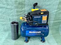 Front angle showing Mastercraft brand name on blue compressor tank and black motor housing, with yellow on/off switch and black hose.