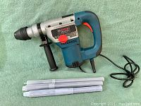 Bosch rotary hammer drill with auxiliary handle and three plastic bit storage tubes on green surface