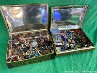 Two large metal tins open and filled with assorted jewelry pieces including bracelets, earrings, necklaces, and brooches.