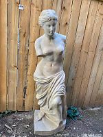 Full view of the life size female sculpture statue against a wooden fence, showing the draped lower body and the base.