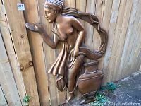 Full view of the large wall sculpture of a goddess showing the whole figure and base against a wooden fence.