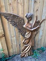 Front view of the large high relief angel sculpture, showing wings, torso, and lyre with a wood fence background.