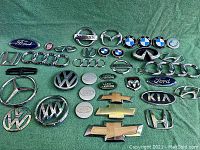 Photo showing a collection of various car manufacturer badges spread out on a green fabric surface, including multiple brands and multiple pieces of some brands.