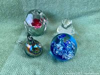 Four vintage art glass paperweights on a green background: faceted clear glass with red rose, clear twisted bubble style, blue with floral design, and iridescent bird shape.