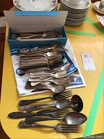 Box and pile of silver plate forks and spoons with some tarnish, laid out on yellow table surface
