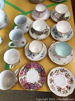 Top-down view of six teacups and saucers with two additional teacups and saucers, showing various floral patterns and gold trim. Cups have different interior colors, including solid white and mint green. Some chipping visible on rims.