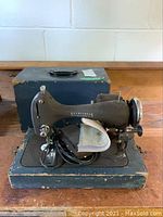 Front view of the National Seamstress sewing machine placed on wooden base with carrying case behind it.