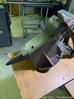 Makeshift anvil with pointed horn and flat mounting base on a wooden surface, showing rust and heavy wear.