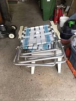 Three stacked metal framed lawn chairs with woven white and blue plastic strap seats and backs, placed on top of a small white plastic garden table.