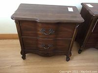 Front and side view of wooden side table showing two drawers with decorative metal handles and carved leg details.
