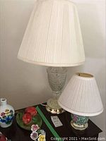 Image showing both lamps: one tall clear etched glass lamp with white pleated shade, and one shorter floral decorated glass lamp with smaller white pleated shade.