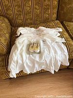 Three embroidered white baby outfits laid out on a couch with one pair of baby shoes on top in packaging.