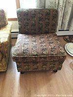 Upholstered armless accent chair with patterned fabric, placed on wooden floor near window with curtains.