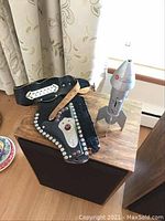 Photo showing the toy cowboy holster with belt and brown wooden toy gun placed on a wooden surface beside the silver metal rocket coin bank.