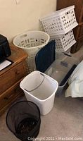 Photo showing large white tall laundry basket, two stacked white plastic laundry baskets, medium transparent plastic storage bin with blue lid, small white rectangular trash bin, and black mesh trash bin arranged near wooden furniture.