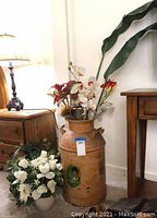 Vintage metal milk jug with painted portrait and faux plants arrangement inside, located near furniture and a lamp