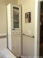 Tall white bathroom cabinet in bathroom corner with folded linen inside glass door, showing two small drawers and lower cabinet door.