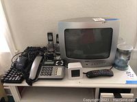 View of vintage RCA CRT TV, Dell keyboard and mouse, Panasonic cordless and wired phones, remote, small device, and water bottle on desk.