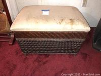 Front view of the rectangular cane storage ottoman chest with stained upholstered top on a red carpet.
