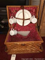 Picnic basket open showing four white enamelware plates attached with leather straps inside the lid along with three enamelware cups hung inside the basket. Basket interior lined with red patterned fabric.