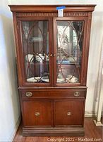 Front view of tall wooden curio cabinet with glass doors featuring wooden oval pattern design. Drawer and bottom cabinet with brass hardware visible.