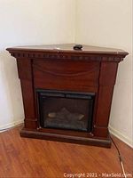 Front view of electric corner fireplace showing wooden mantel and firebox with faux logs. Power cord visible on floor. Remote control and card on mantel.