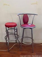 Two metal counter stools with red cushioned seats on a hardwood floor against a beige wall; one stool has a backrest, the other does not.