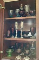 Full view inside wooden cabinet showing multiple shelves filled with tall goblets, stemmed glasses, various tumblers, and bowls in purple, clear and green glass.