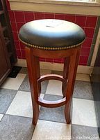 Side view of wooden counter stool showing wood grain, four legs, circular footrest, and leather cushion top with nailhead trim.