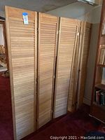 Four wooden shutter doors made with horizontal slats, standing upright in room, showing natural wood finish and metal hinges connecting panels.