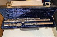 Open black hard case lined with dark blue velvet showing disassembled silver-colored metal flute in three parts: footjoint, body, and headjoint.