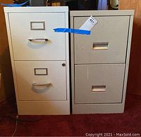 Front view of two vertical metal file cabinets placed side by side on carpet. One cabinet beige with three drawers, second cabinet light gray with two drawers.