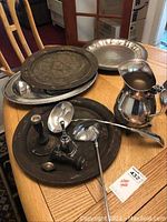 Wide shot showing all items in the lot grouped on a wooden table: includes large footed platter, pitcher, ladles, pierced tray, candle holders and serving utensils.