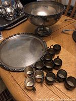 View of the punch bowl, underplate, and 12 cups, two cups missing handles, showing tarnish and wear