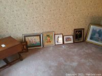 Seven framed artworks lined up on carpet against a floral wall, includes various sizes, one frame visibly damaged