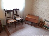 Photo showing two wooden caned chairs and a wooden low drop leaf table against wall with floral wallpaper, carpeted floor. One chair has caning damage.
