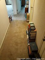 View down hallway showing stacks of hardcover books on carpeted floor along walls, with a small wooden bookcase containing encyclopedias.