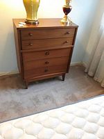 Front view of the mid century dresser showing five drawers; three have concave fronts with rectangular handles, two have straight fronts with round knobs. The dresser is on a carpeted floor beside a bed with lamps on top.