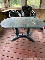 Oval patio table with marble-look plastic top showing scratches and edge damage along with green plastic chairs stacked behind it.
