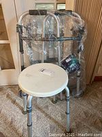 Photo of silver Invacare walker wrapped in plastic behind white circular shower stool showing details of the lot