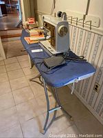 Singer Touch & Sew model 620 sewing machine placed on blue covered metal frame ironing board in laundry room, showing metal foot pedal and multiple accessories including thread spools and manuals.