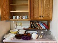 Overview of countertop with various kitchen items including CorningWare baking dishes, Pyrex bowl, cookbooks and mugs on shelves above.