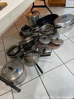 Photo of assorted aluminum pots, pans, roasters, and lids on white tiled floor.