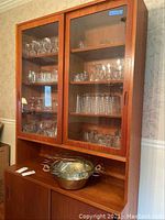 Glass cabinet containing assorted stemware and glassware including goblets, wine glasses, juice glasses, shot glasses, and bowls, with metal serving bowl and wicker handles placed beneath.