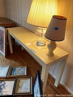 Full view of the white laminated sofa table in a living space with lamps on top and framed pictures underneath.