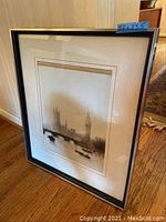 Framed wall art depicting Westminster landmarks on hardwood floor, angled view showing reflection in glass.